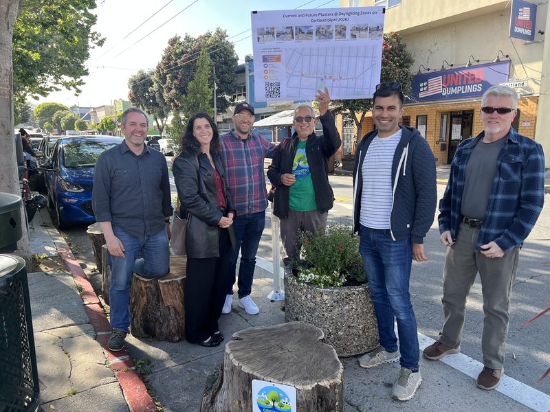 Six neighbors smile beside stone planters and log seats at the Cortland Avenue pilot corner in Bernal Heights.