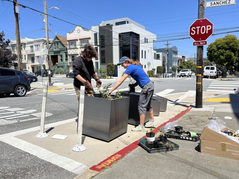 Two volunteers set up steel planters beside white bollards at the Sanchez Street corner in Noe Valley.