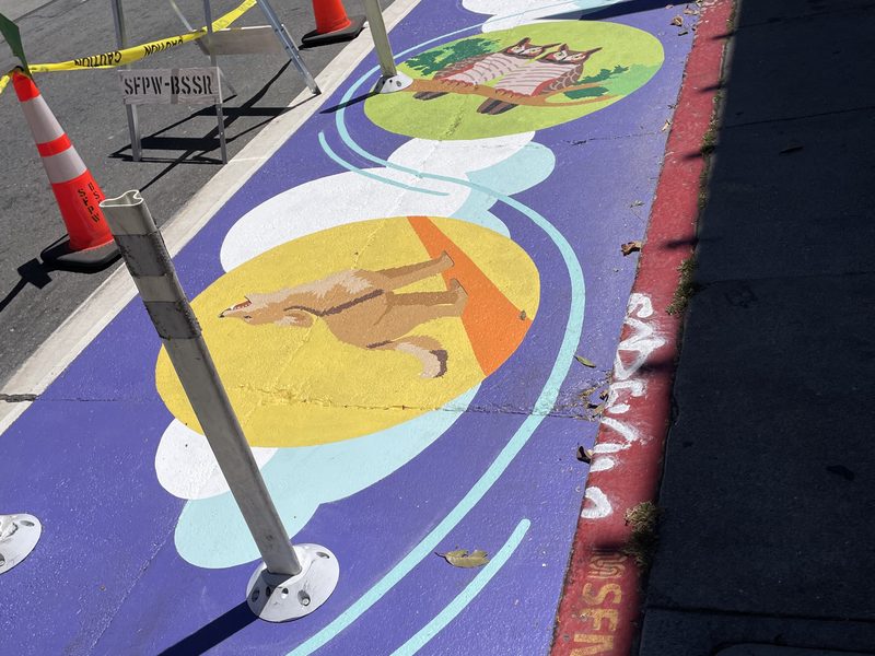 A freshly painted purple street mural with round medallions of a coyote and an owl runs along the curb inside a safety zone.