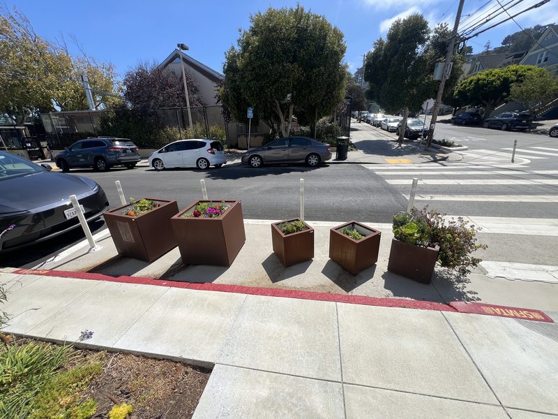 Three rust-colored steel planters filled with flowers sit inside a painted safety zone at a San Francisco corner.