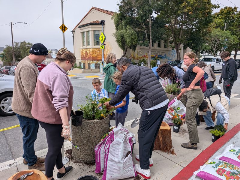 Neighbors of all ages, including kids, fill stone planters with flowers and potting soil at a corner on planting day.