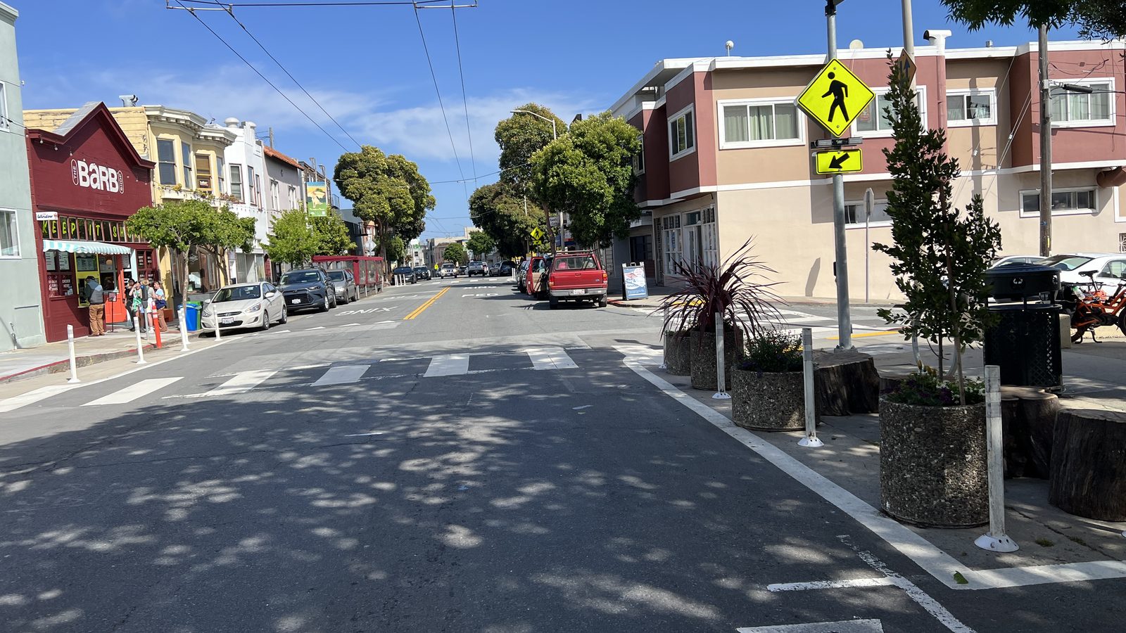 The Cortland Avenue Community Corner in Bernal Heights: stone planters and log seats brighten a daylit corner beside a crosswalk on a sunny afternoon.