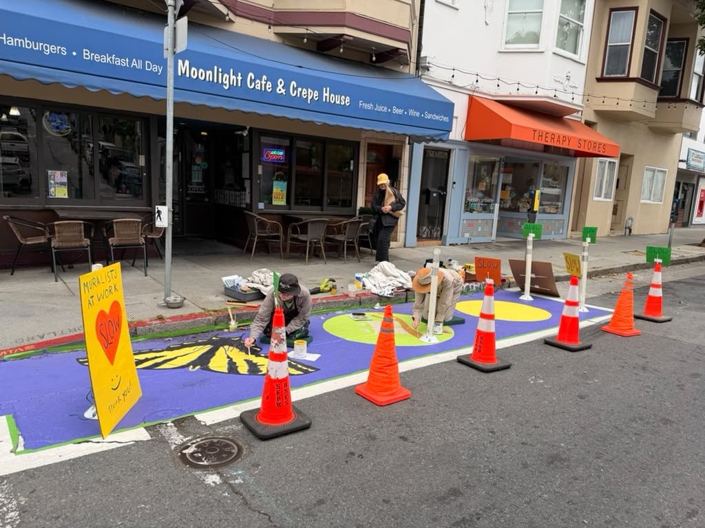 Artists kneel on the street painting a purple and yellow mural inside a coned-off safety zone in front of a neighborhood cafe.